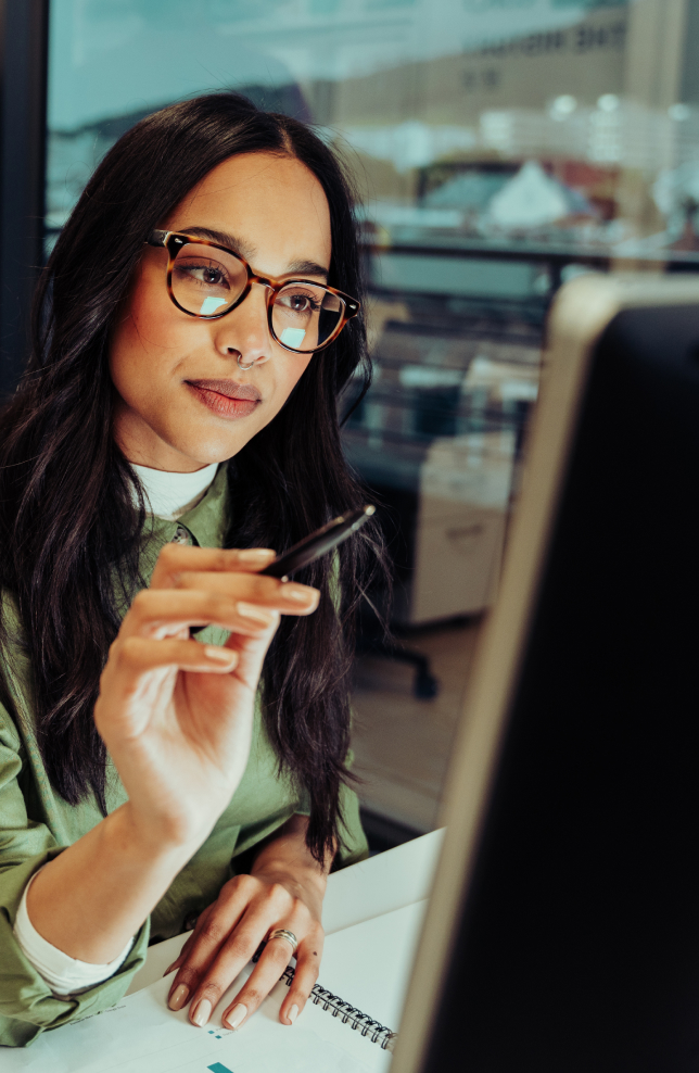 Woman with glasses holding pen looking at desktop screen